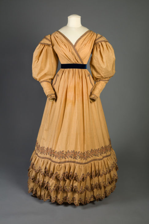 Tan/yellow muslin dress with three tiers of ruffles at hem and gigot sleeves.