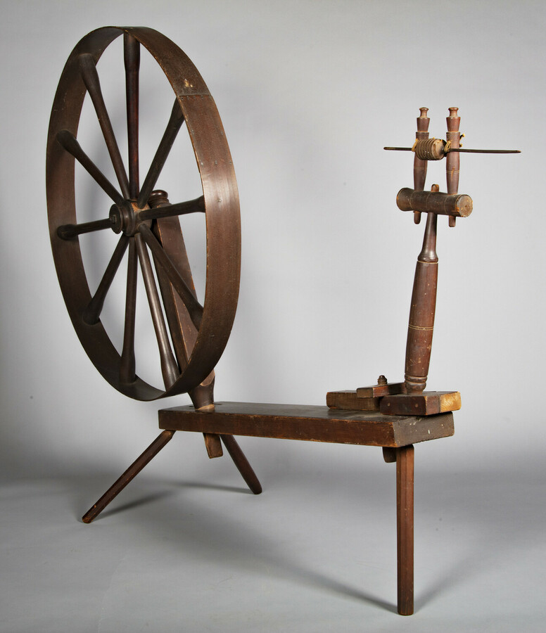 Image of a wooden flax spinning wheel with the wheel on the left side and the spindle on the right on a gray background.