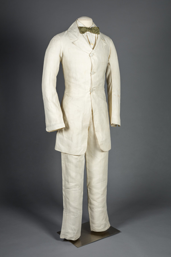 A white linen suit comprised of a jacket, pants, and a shirt worn by a mannequin with a patterned bow tie. Image has gray background.