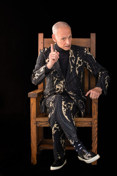 Photograph of John Waters sitting in a chair.