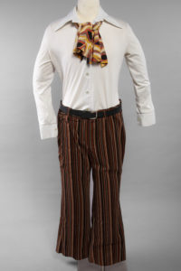 Image of a mannequin dressed in a white collared shirt worn with an orange, yellow, and brown ascot, and orange, yellow, and brown stripped corduroy pants worn with a black belt.