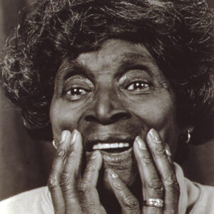 Black and white portrait of Scott smiling, both hands are held to her lower face. Her face and hands fill the frame.