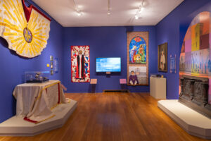 View of three walls from the exhibition "Stitched in Favor". The gallery is painted a rich purple, colorful panels, quilts, and artwork adorn the walls.
