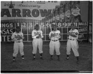 Group portrait of four batters of the Elite Giants, a Negro League baseball team from Baltimore, Maryland. Left to right: Henry Kimbo, Robert "Butch" Davis, Lester Locket, and Lenny Pearson. Behind them is a scoreboard and advertisement celebrating the 200th anniversary of Arrow Beer.