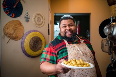 A photograph of a man smiling and holding a plate of food. He is dressed in a red, green, and black shirt with a white apron on, and he is standing in a kitchen with pots and bowls hanging on the wall behind him.